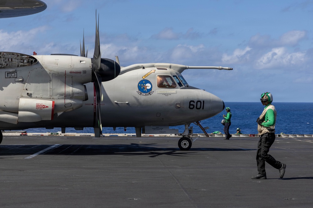 A U.S. Navy E-2 Hawkeye airborne early warning aircraft, numbered 601, is positioned on the flight deck of an aircraft carrier at sea. Deck crew members wearing green jerseys and helmets are visible moving around the aircraft. The scene depicts routine flight deck operations under clear blue skies over open ocean.