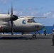 A U.S. Navy E-2 Hawkeye airborne early warning aircraft, numbered 601, is positioned on the flight deck of an aircraft carrier at sea. Deck crew members wearing green jerseys and helmets are visible moving around the aircraft. The scene depicts routine flight deck operations under clear blue skies over open ocean.