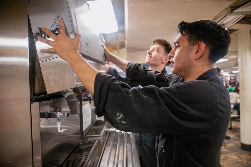 Two young men in dark work uniforms are collaborating to install or adjust a panel on a stainless steel commercial beverage or food service machine. The setting appears to be a large institutional kitchen or cafeteria environment. The foreground worker is of Asian descent and the background worker appears Caucasian, both focused on the task.