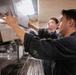 Two young men in dark work uniforms are collaborating to install or adjust a panel on a stainless steel commercial beverage or food service machine. The setting appears to be a large institutional kitchen or cafeteria environment. The foreground worker is of Asian descent and the background worker appears Caucasian, both focused on the task.