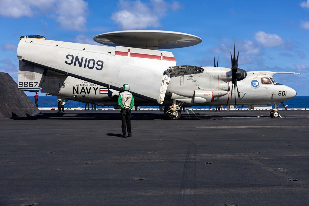 A U.S. Navy E-2D Advanced Hawkeye airborne early warning aircraft (bureau number 9867, modex 601) is parked on the flight deck of USS Abraham Lincoln (CVN-72), assigned to VAW-117. The aircraft's wings are folded for carrier deck storage, and its distinctive rotodome radar disc is clearly visible atop the fuselage. A flight deck crew member in a green jersey stands in the foreground observing the aircraft.