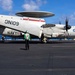 A U.S. Navy E-2D Advanced Hawkeye airborne early warning aircraft (bureau number 9867, modex 601) is parked on the flight deck of USS Abraham Lincoln (CVN-72), assigned to VAW-117. The aircraft's wings are folded for carrier deck storage, and its distinctive rotodome radar disc is clearly visible atop the fuselage. A flight deck crew member in a green jersey stands in the foreground observing the aircraft.