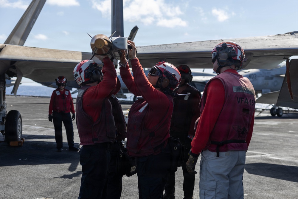Navy ordnance personnel wearing red jerseys and helmets are loading or attaching a missile to the wing pylon of a carrier-based aircraft on a flight deck. The crew members are identifiable as belonging to VFA-15 squadron based on visible vest markings. The scene takes place on what appears to be a U.S. Navy aircraft carrier flight deck under partly cloudy skies.