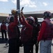 Navy ordnance personnel wearing red jerseys and helmets are loading or attaching a missile to the wing pylon of a carrier-based aircraft on a flight deck. The crew members are identifiable as belonging to VFA-15 squadron based on visible vest markings. The scene takes place on what appears to be a U.S. Navy aircraft carrier flight deck under partly cloudy skies.