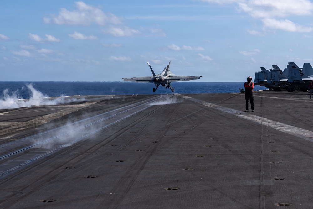A carrier-based jet aircraft, consistent with an F/A-18 Hornet or Super Hornet, is being catapult-launched from the flight deck of a US Navy aircraft carrier at sea. Steam from the catapult system is visible trailing behind the aircraft as it lifts off the deck. A flight deck crew member in a red jersey stands observing the launch, with additional parked aircraft visible on the right side of the deck.