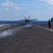 A carrier-based jet aircraft, consistent with an F/A-18 Hornet or Super Hornet, is being catapult-launched from the flight deck of a US Navy aircraft carrier at sea. Steam from the catapult system is visible trailing behind the aircraft as it lifts off the deck. A flight deck crew member in a red jersey stands observing the launch, with additional parked aircraft visible on the right side of the deck.
