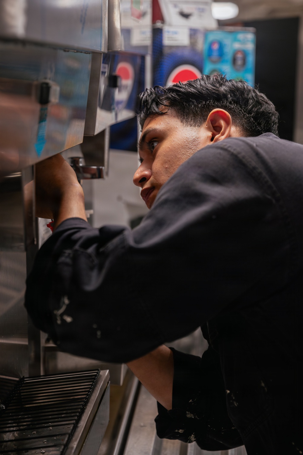 A young man wearing a dark uniform is leaning in closely to inspect or service a stainless steel soda fountain or beverage dispensing machine. The setting appears to be a commercial food service environment, likely a fast food restaurant or cafeteria. A Pepsi branded display or machine is visible in the background.