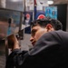 A young man wearing a dark uniform is leaning in closely to inspect or service a stainless steel soda fountain or beverage dispensing machine. The setting appears to be a commercial food service environment, likely a fast food restaurant or cafeteria. A Pepsi branded display or machine is visible in the background.