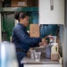 A woman wearing a navy blue uniform is working behind a counter preparing beverages. The setting appears to be a small Starbucks kiosk or coffee station, likely aboard a military vessel or installation based on the industrial green-painted metal walls and the worker's uniform. Two Starbucks clear plastic cups are visible on the stainless steel counter along with beverage supplies.
