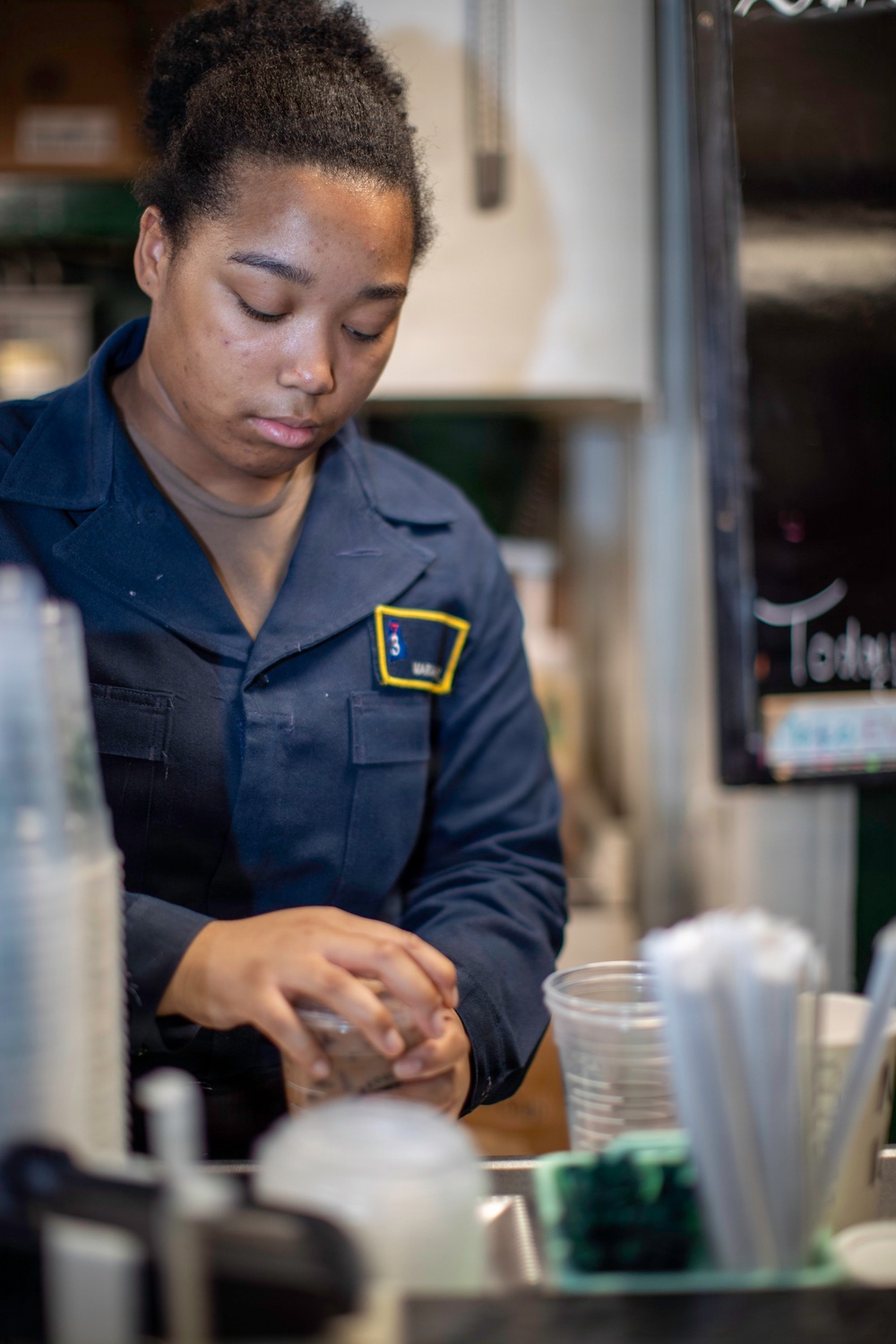 A young woman wearing a dark navy uniform with a yellow-bordered patch/insignia is working behind a service counter. She appears to be handling cups or containers in what looks like a food or beverage service setting. The environment suggests a cafeteria, canteen, or similar service establishment.