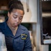 A young woman wearing a dark navy uniform with a yellow-bordered patch/insignia is working behind a service counter. She appears to be handling cups or containers in what looks like a food or beverage service setting. The environment suggests a cafeteria, canteen, or similar service establishment.