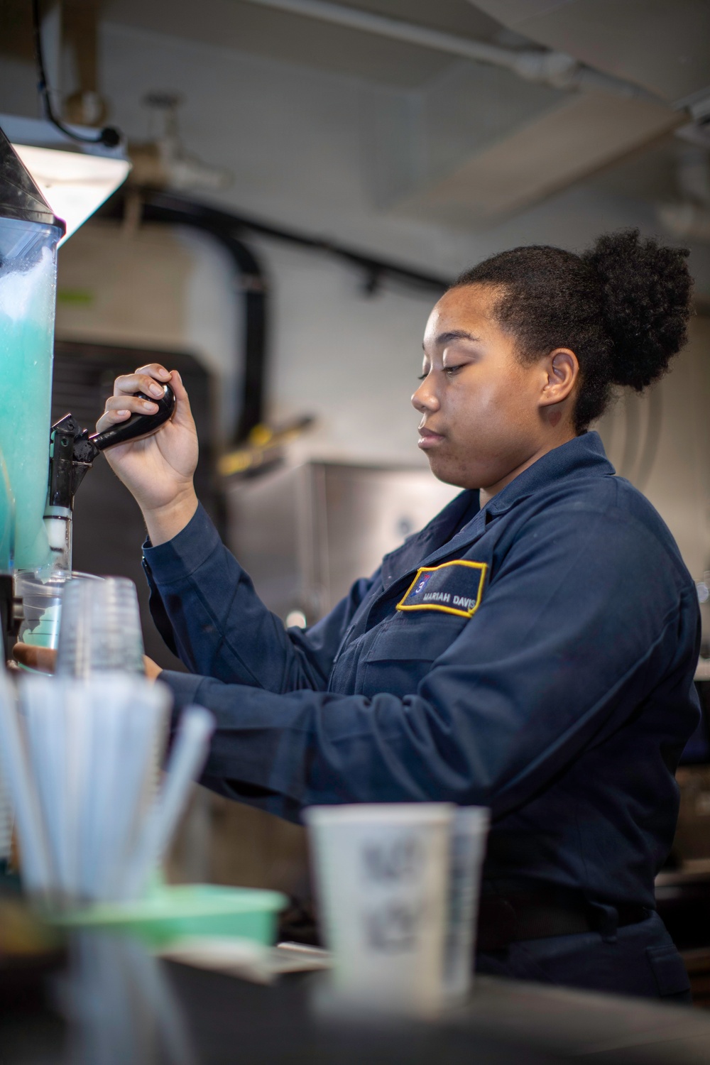 A person wearing a dark navy blue uniform with a name patch reading 'MARIAH DAVIS' is operating what appears to be a beverage dispensing machine, likely a slushie or frozen drink machine containing a teal/blue liquid. The setting appears to be a military or institutional food service environment, possibly aboard a naval vessel given the industrial ceiling infrastructure visible in the background. The individual is focused on the task, handling a dispenser handle with stacked plastic cups and straws visible in the foreground.