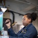 A person wearing a dark navy blue uniform with a name patch reading 'MARIAH DAVIS' is operating what appears to be a beverage dispensing machine, likely a slushie or frozen drink machine containing a teal/blue liquid. The setting appears to be a military or institutional food service environment, possibly aboard a naval vessel given the industrial ceiling infrastructure visible in the background. The individual is focused on the task, handling a dispenser handle with stacked plastic cups and straws visible in the foreground.