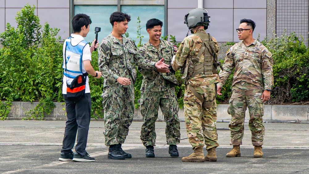 A group of military personnel from different branches are gathered outdoors on a paved surface, appearing to interact and shake hands. Two individuals are wearing Japanese Maritime Self-Defense Force (JMSDF) digital camouflage uniforms, while two others wear U.S. Army MultiCam (OCP) uniforms, one of whom is wearing a flight helmet. A civilian in a blue and white vest is holding a camera on a selfie stick, suggesting media coverage or documentation of the event.