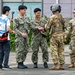 A group of military personnel from different branches are gathered outdoors on a paved surface, appearing to interact and shake hands. Two individuals are wearing Japanese Maritime Self-Defense Force (JMSDF) digital camouflage uniforms, while two others wear U.S. Army MultiCam (OCP) uniforms, one of whom is wearing a flight helmet. A civilian in a blue and white vest is holding a camera on a selfie stick, suggesting media coverage or documentation of the event.