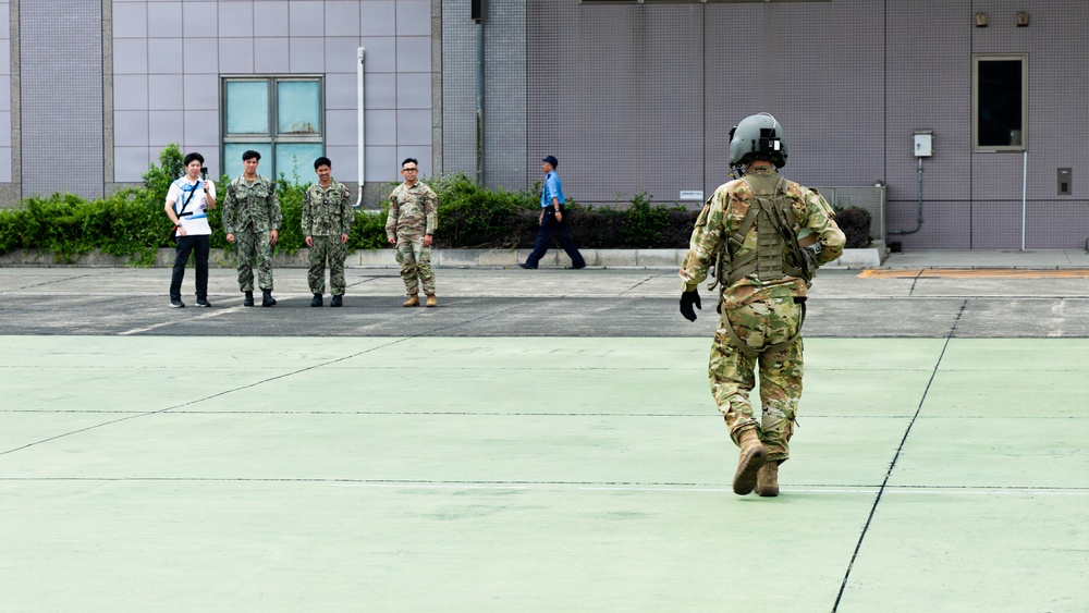 The image shows a military tarmac or helipad area where personnel in various uniforms are present. A soldier in full combat gear including a flight helmet and multicam/OCP camouflage is walking across the green-painted surface toward a group of individuals. The group in the background includes personnel in what appear to be navy/maritime camouflage uniforms, one person in tan/army camouflage, a civilian in white, and a person in a blue uniform.