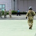 The image shows a military tarmac or helipad area where personnel in various uniforms are present. A soldier in full combat gear including a flight helmet and multicam/OCP camouflage is walking across the green-painted surface toward a group of individuals. The group in the background includes personnel in what appear to be navy/maritime camouflage uniforms, one person in tan/army camouflage, a civilian in white, and a person in a blue uniform.