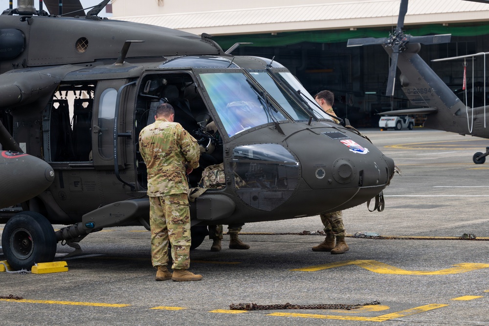 Military personnel in camouflage uniforms are conducting maintenance or pre-flight checks on a dark olive-green UH-60 Black Hawk helicopter on a tarmac. At least three soldiers are visible working around the aircraft's cockpit and cabin area. The setting appears to be a military airfield with a hangar visible in the background.