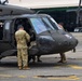 Military personnel in camouflage uniforms are conducting maintenance or pre-flight checks on a dark olive-green UH-60 Black Hawk helicopter on a tarmac. At least three soldiers are visible working around the aircraft's cockpit and cabin area. The setting appears to be a military airfield with a hangar visible in the background.