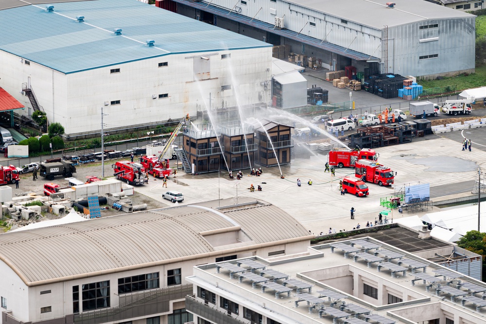 An aerial view of what appears to be a firefighting drill or training exercise at an industrial facility in Japan. Multiple red fire trucks are deployed around a small multi-story structure, with water streams being directed at the building from several angles including an aerial ladder truck. Numerous personnel in firefighting gear and observers are visible throughout the scene.
