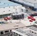 An aerial view of what appears to be a firefighting drill or training exercise at an industrial facility in Japan. Multiple red fire trucks are deployed around a small multi-story structure, with water streams being directed at the building from several angles including an aerial ladder truck. Numerous personnel in firefighting gear and observers are visible throughout the scene.