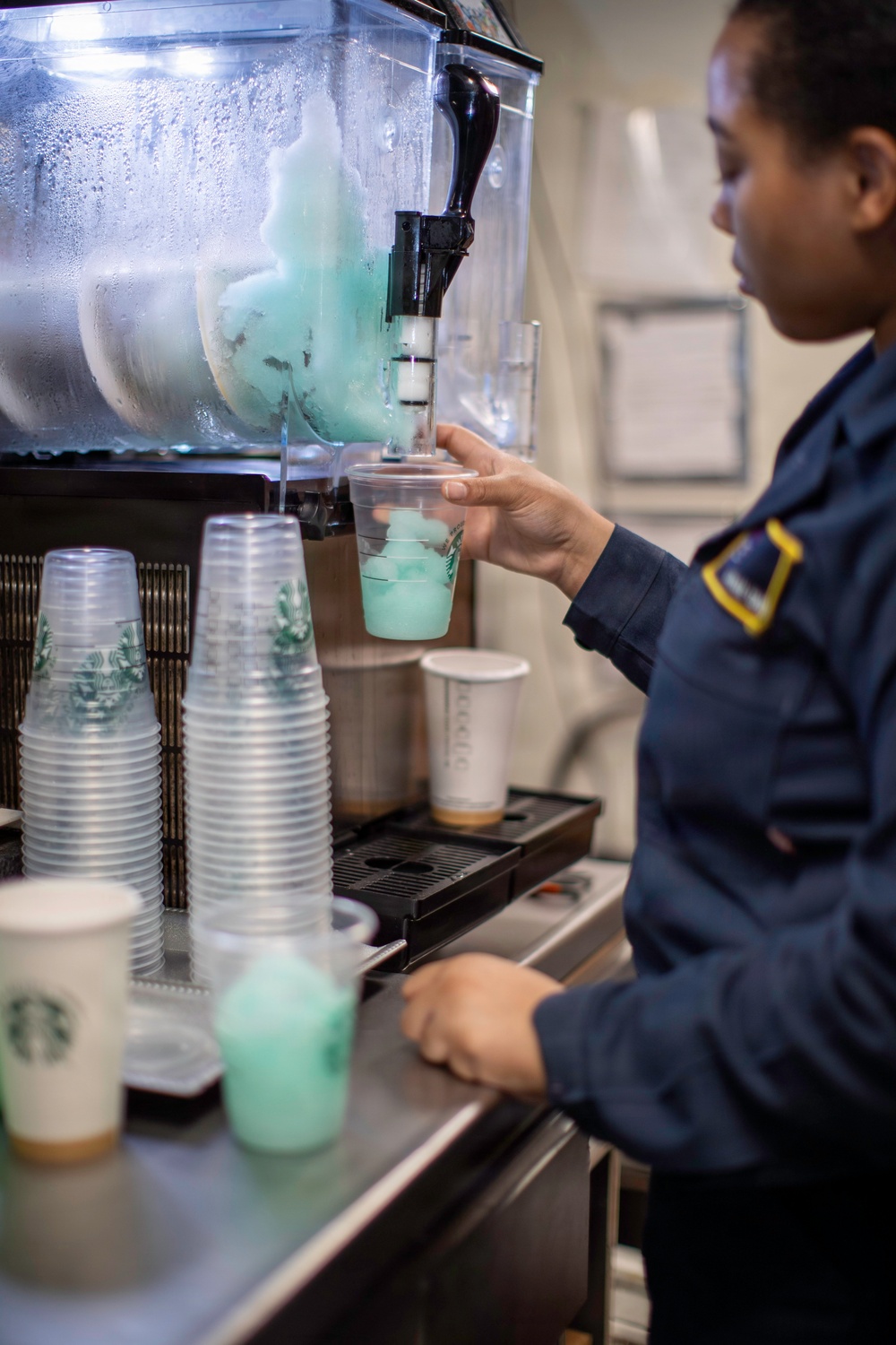 A person in a dark navy uniform with a yellow shoulder patch is filling a clear plastic cup with a teal/mint-colored frozen slush beverage from a commercial slush machine. The setting appears to be a Starbucks service counter, evidenced by branded cups visible in the foreground. Multiple stacked clear plastic cups and a Starbucks paper cup are visible on the counter.