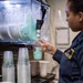 A person in a dark navy uniform with a yellow shoulder patch is filling a clear plastic cup with a teal/mint-colored frozen slush beverage from a commercial slush machine. The setting appears to be a Starbucks service counter, evidenced by branded cups visible in the foreground. Multiple stacked clear plastic cups and a Starbucks paper cup are visible on the counter.
