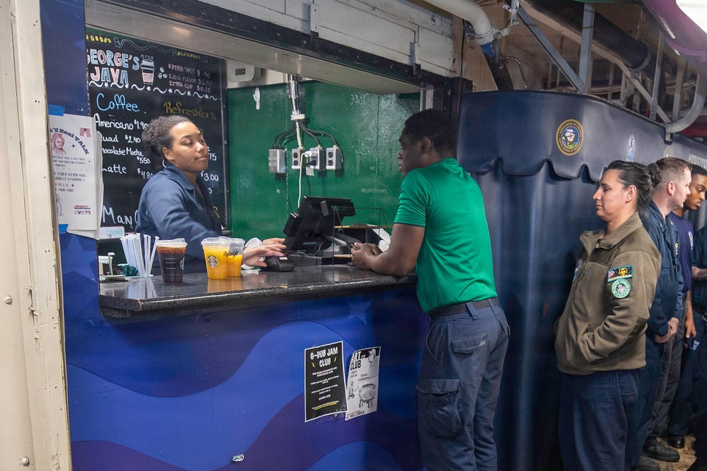 The image shows a coffee shop called 'George's Java' operating aboard what appears to be a U.S. Navy aircraft carrier, identifiable by the naval insignia and industrial surroundings. A female barista is serving customers at the counter, with several personnel in military-style uniforms waiting in line. Starbucks-branded cups are visible on the counter, suggesting a licensed or affiliated coffee operation.