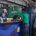 The image shows a coffee shop called 'George's Java' operating aboard what appears to be a U.S. Navy aircraft carrier, identifiable by the naval insignia and industrial surroundings. A female barista is serving customers at the counter, with several personnel in military-style uniforms waiting in line. Starbucks-branded cups are visible on the counter, suggesting a licensed or affiliated coffee operation.