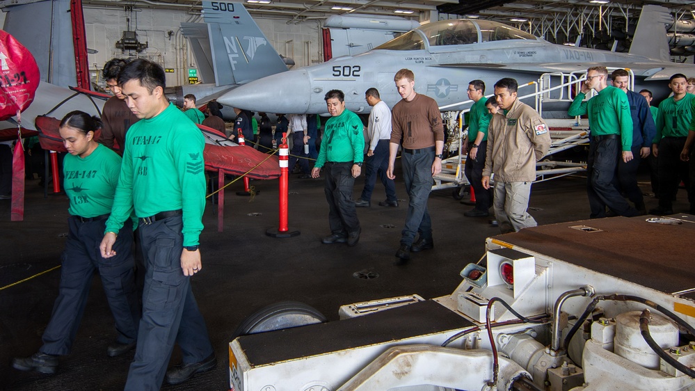 The image shows U.S. Navy personnel conducting a FOD (Foreign Object Debris) walkdown in the hangar bay of an aircraft carrier. Multiple sailors wearing green jerseys marked 'VTAA-147' are walking in a line, scanning the deck for debris. Military aircraft, including jets marked '502' and '500' with 'VAQ-141' squadron markings, are visible in the background.