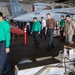 The image shows U.S. Navy personnel conducting a FOD (Foreign Object Debris) walkdown in the hangar bay of an aircraft carrier. Multiple sailors wearing green jerseys marked 'VTAA-147' are walking in a line, scanning the deck for debris. Military aircraft, including jets marked '502' and '500' with 'VAQ-141' squadron markings, are visible in the background.