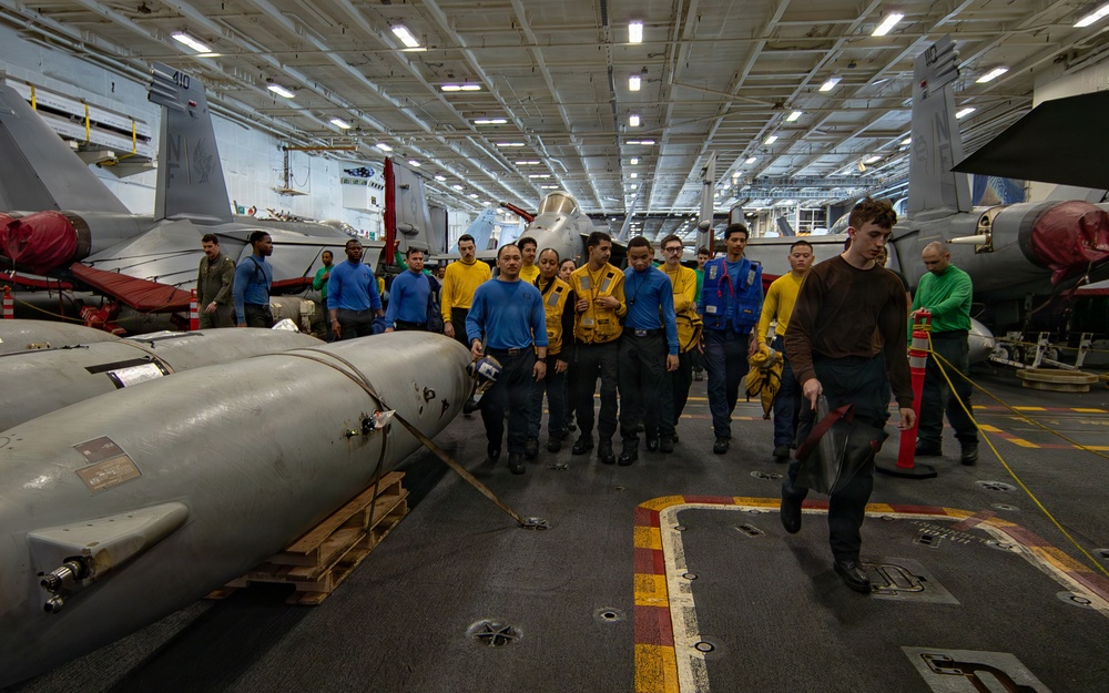 The image shows a group of U.S. Navy personnel gathered in the hangar bay of an aircraft carrier, surrounded by military aircraft and ordnance. Personnel are wearing color-coded jerseys (yellow, blue, brown, green) typical of carrier flight deck and hangar deck crew designations. A large munition or external fuel tank is visible on a wooden pallet in the foreground.