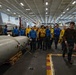 The image shows a group of U.S. Navy personnel gathered in the hangar bay of an aircraft carrier, surrounded by military aircraft and ordnance. Personnel are wearing color-coded jerseys (yellow, blue, brown, green) typical of carrier flight deck and hangar deck crew designations. A large munition or external fuel tank is visible on a wooden pallet in the foreground.