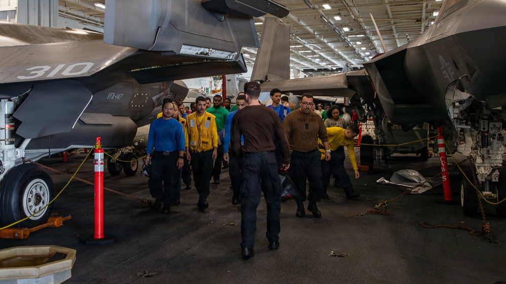 The image shows a group of U.S. Navy personnel walking through an aircraft carrier hangar bay between two F-35C Lightning II fighter jets. Crew members are wearing color-coded jerseys indicating their roles, consistent with standard carrier air wing operations. The setting appears to be an active hangar deck, likely aboard a Nimitz or Gerald R. Ford-class carrier.