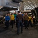 The image shows a group of U.S. Navy personnel walking through an aircraft carrier hangar bay between two F-35C Lightning II fighter jets. Crew members are wearing color-coded jerseys indicating their roles, consistent with standard carrier air wing operations. The setting appears to be an active hangar deck, likely aboard a Nimitz or Gerald R. Ford-class carrier.