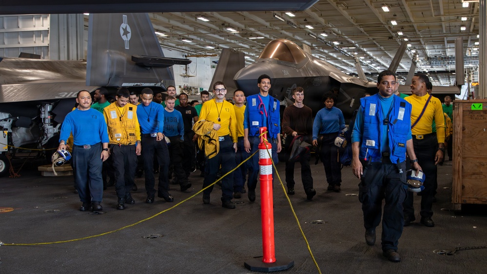 The image shows a group of U.S. Navy personnel walking through an aircraft carrier hangar bay. Multiple crew members are wearing color-coded jerseys and vests in blue and yellow, which indicate their specific roles in flight deck operations. In the background, at least two F-35C Lightning II stealth fighter jets are visible, parked and secured in the hangar.