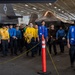 The image shows a group of U.S. Navy personnel walking through an aircraft carrier hangar bay. Multiple crew members are wearing color-coded jerseys and vests in blue and yellow, which indicate their specific roles in flight deck operations. In the background, at least two F-35C Lightning II stealth fighter jets are visible, parked and secured in the hangar.