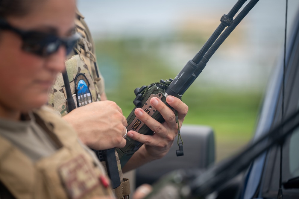 A military or law enforcement personnel in MultiCam/OCP camouflage tactical gear is operating a handheld radio device, likely a military-grade communications unit. The individual is wearing sunglasses and a tactical vest with a visible unit patch on the shoulder. The setting appears to be outdoors near a vehicle.