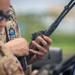 A military or law enforcement personnel in MultiCam/OCP camouflage tactical gear is operating a handheld radio device, likely a military-grade communications unit. The individual is wearing sunglasses and a tactical vest with a visible unit patch on the shoulder. The setting appears to be outdoors near a vehicle.