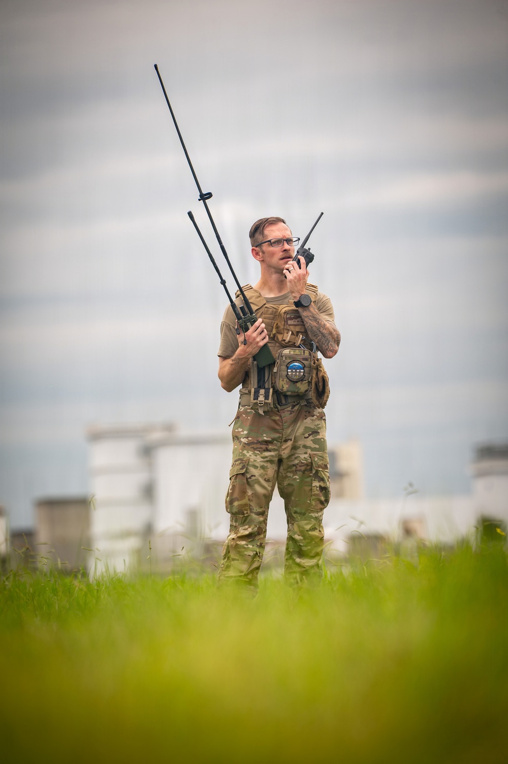 A male individual in military-style multicam camouflage uniform and tactical vest stands in an open grassy field, operating handheld radio communications equipment. He is holding what appears to be a military radio with multiple extended antennas and speaking into a handheld transceiver. White industrial or military structures are visible in the blurred background.