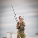 A male individual in military-style multicam camouflage uniform and tactical vest stands in an open grassy field, operating handheld radio communications equipment. He is holding what appears to be a military radio with multiple extended antennas and speaking into a handheld transceiver. White industrial or military structures are visible in the blurred background.