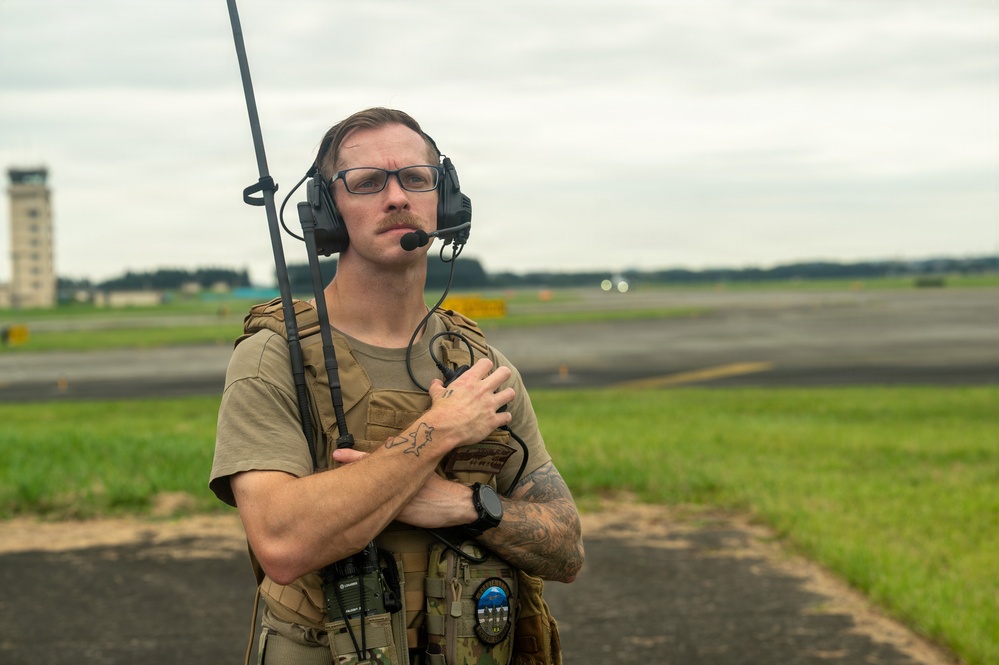 A military or security personnel member stands on an airfield tarmac wearing tactical gear including a plate carrier vest and communications headset with a radio antenna. He appears to be monitoring or coordinating operations on the flight line, with an air traffic control tower visible in the background. The subject has tattoos on his forearms, wears glasses, and has a mustache.