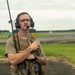 A military or security personnel member stands on an airfield tarmac wearing tactical gear including a plate carrier vest and communications headset with a radio antenna. He appears to be monitoring or coordinating operations on the flight line, with an air traffic control tower visible in the background. The subject has tattoos on his forearms, wears glasses, and has a mustache.