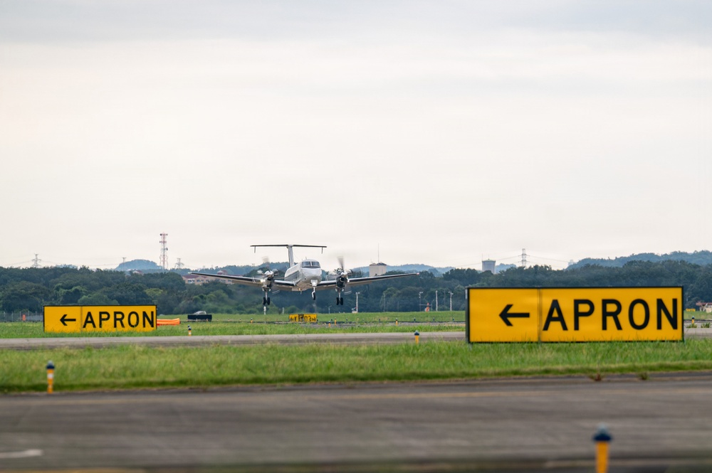 A twin-engine turboprop aircraft is taxiing or landing on a runway at an airport. Two yellow 'APRON' directional signs with left-pointing arrows are prominently visible in the foreground on either side of the taxiway. The background shows a tree line, communication towers, and an overcast sky.