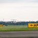 A twin-engine turboprop aircraft is taxiing or landing on a runway at an airport. Two yellow 'APRON' directional signs with left-pointing arrows are prominently visible in the foreground on either side of the taxiway. The background shows a tree line, communication towers, and an overcast sky.