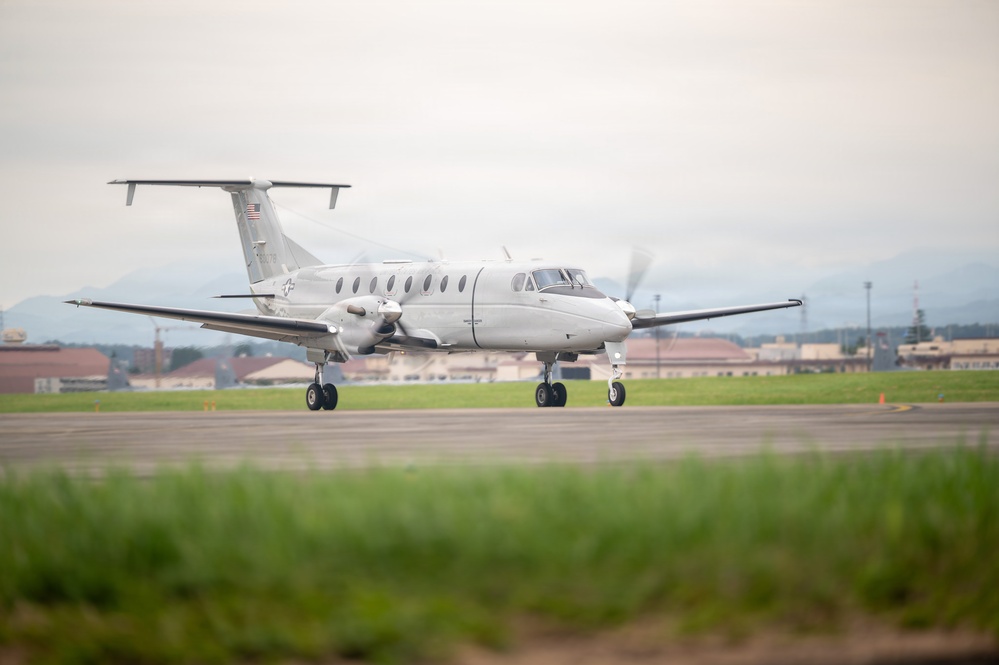 A white twin-turboprop military aircraft is taxiing on a runway at what appears to be a military airfield. The aircraft displays a U.S. flag on its tail and military insignia, consistent with a Beechcraft C-12 Huron or similar platform. Mountains and military base infrastructure are visible in the background.