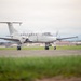 A white twin-turboprop military aircraft is taxiing on a runway at what appears to be a military airfield. The aircraft displays a U.S. flag on its tail and military insignia, consistent with a Beechcraft C-12 Huron or similar platform. Mountains and military base infrastructure are visible in the background.