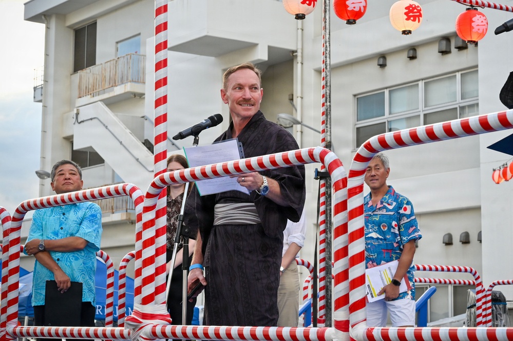 A man wearing a traditional Japanese yukata (dark gray) is speaking at a microphone on a decorated stage, holding papers and smiling. The setting appears to be a Japanese festival (matsuri), indicated by red and white striped decorations and Japanese paper lanterns with the character '祭' (festival) visible in the background. Several other individuals stand behind him on the stage, including two men in colorful Hawaiian-style shirts.