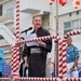 A man wearing a traditional Japanese yukata (dark gray) is speaking at a microphone on a decorated stage, holding papers and smiling. The setting appears to be a Japanese festival (matsuri), indicated by red and white striped decorations and Japanese paper lanterns with the character '祭' (festival) visible in the background. Several other individuals stand behind him on the stage, including two men in colorful Hawaiian-style shirts.
