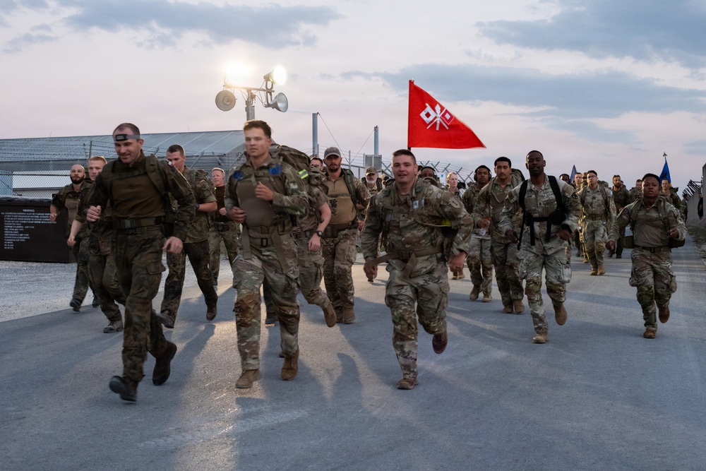 A large group of military personnel in camouflage uniforms are conducting a formation run or ruck march on a paved surface during early morning or dusk hours. A red guidon flag with a white crossed-keys or signal insignia is visible being carried mid-formation. The setting appears to be a military installation or base, with floodlights, fencing, and industrial structures visible in the background.